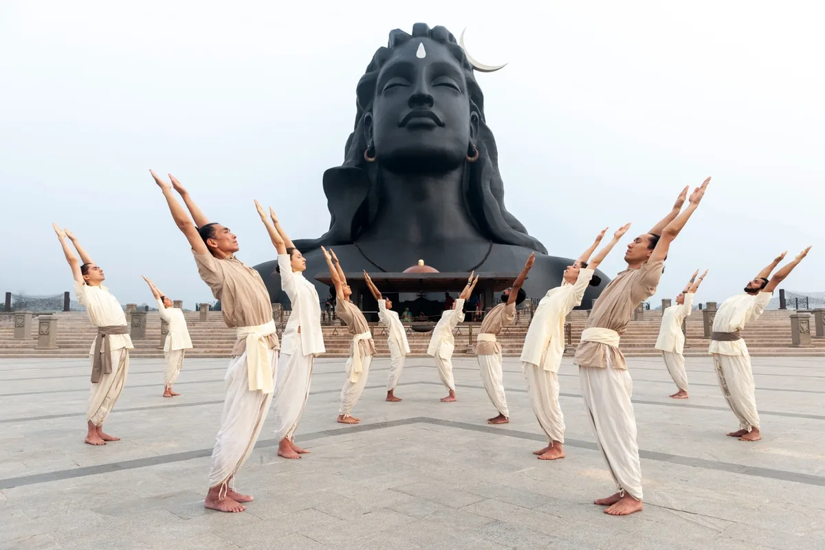 Praticanti di Hatha Yoga vestiti di bianco, con le braccia alzate, davanti al grande volto di Adiyogi